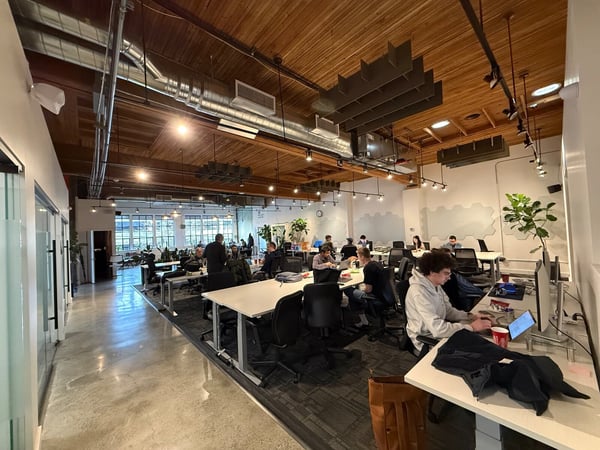 A wide-angle photograph captures a modern, open-plan office environment with high wooden ceilings and exposed industrial elements, where several people are seated at desks working on computers. The space appears well-lit, combining white walls with warm wood tones and concrete flooring.