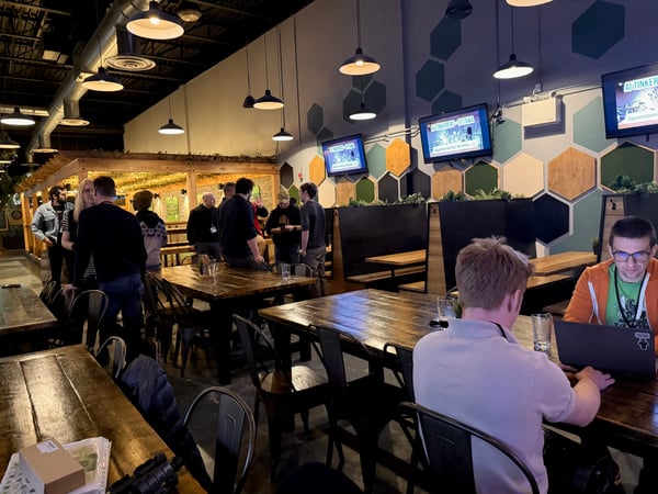 A photo of an indoor social gathering or event in a modern space with wooden tables, industrial lighting, and people networking.