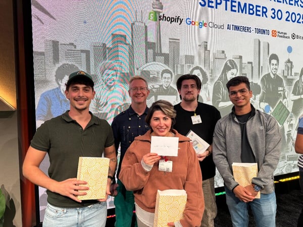 A group of five smiling individuals, likely attendees or participants, pose for a photo at an indoor event, holding wrapped gifts or prizes in front of a large digital screen displaying sponsor logos and event information.