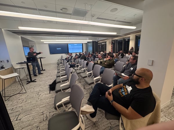 A photograph captures an indoor professional presentation taking place in a modern conference room, showing a male presenter standing near two large screens addressing a seated audience of approximately thirty people.