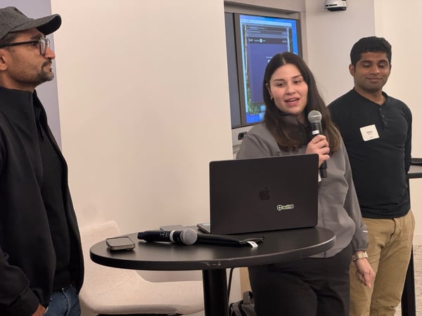 A candid photo captures three people indoors during a technical presentation, with a woman in the center speaking into a microphone while standing next to a black laptop.