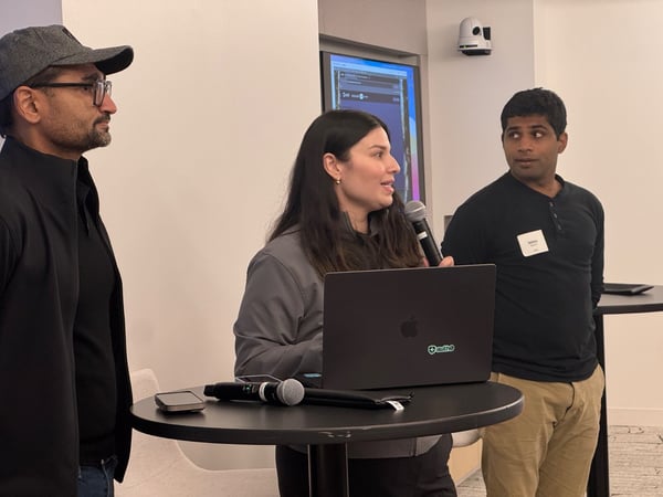 A candid photograph captures three people—a woman speaking into a microphone and two men flanking her—participating in an indoor presentation or discussion, with an open laptop resting on a small table.
