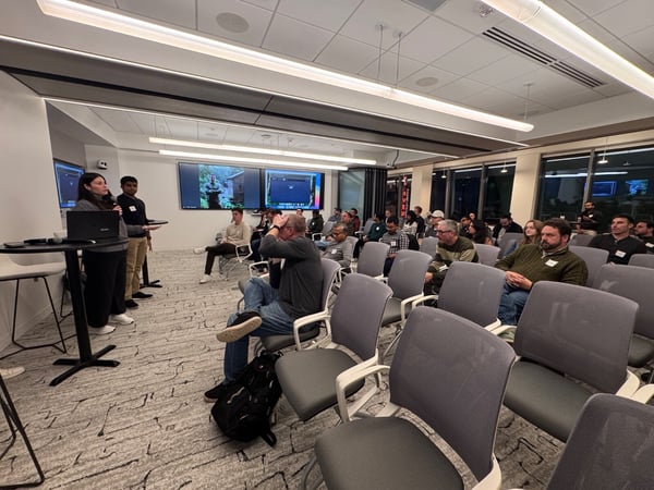 A photograph captures an indoor presentation where two speakers stand at the front, addressing a large seated audience in a modern conference room with multiple large screens displaying content.