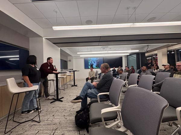A photograph capturing an indoor presentation or discussion event, showing two speakers standing near a counter addressing a large, seated audience in a modern conference room.