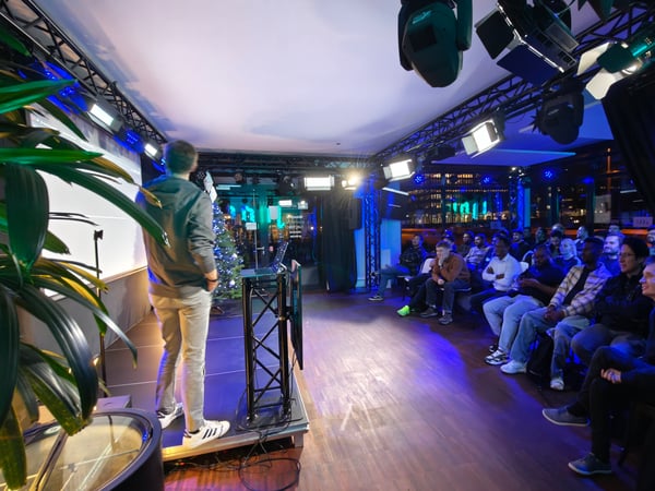 A man is giving a presentation on a stage to a seated audience in a dimly lit room with blue and white stage lighting.
