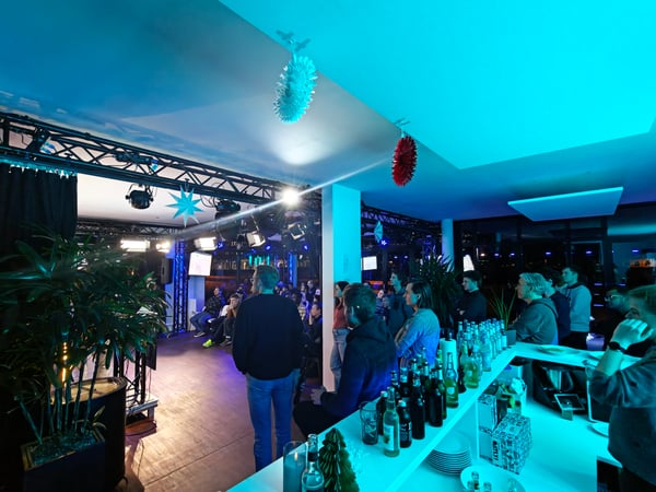 A wide-angle photograph of an indoor event space where a crowd is gathered for a presentation or demo, featuring a bar in the foreground and vibrant blue and purple lighting.