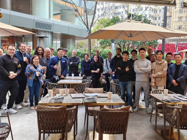 A large group of approximately 23 people, likely colleagues, pose for a group photo on an outdoor urban patio featuring tables, chairs, and festive red Chinese New Year decorations.