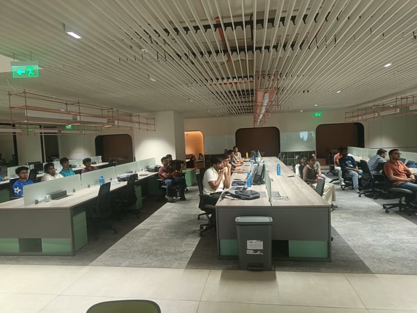 A group of people are seated at long, modern desks in a large office or coworking space, appearing to attend a presentation or meeting.