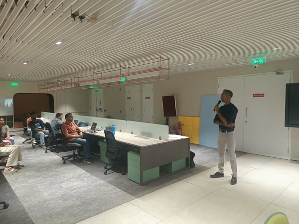 A man stands in a modern office, speaking into a microphone while a small group of people sits at desks listening to him.