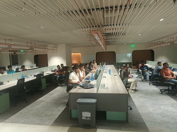 A group of people are seated at long communal desks in a modern, open-plan office space, attentively watching a presentation or demonstration.