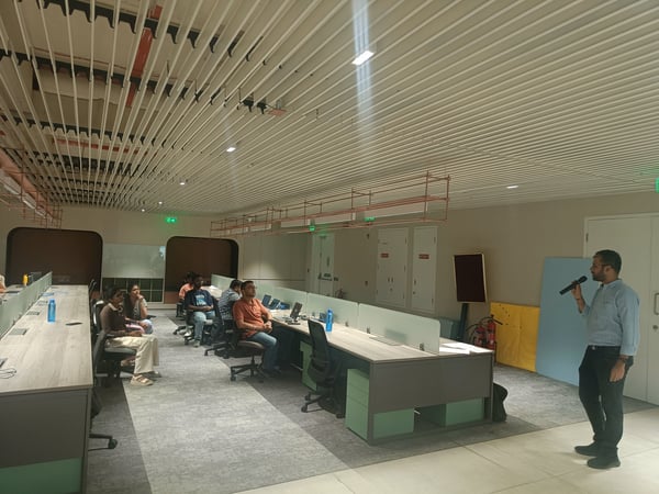 A man is giving a presentation with a microphone in a modern, open-plan office. A small group of people is seated at desks, listening to him.