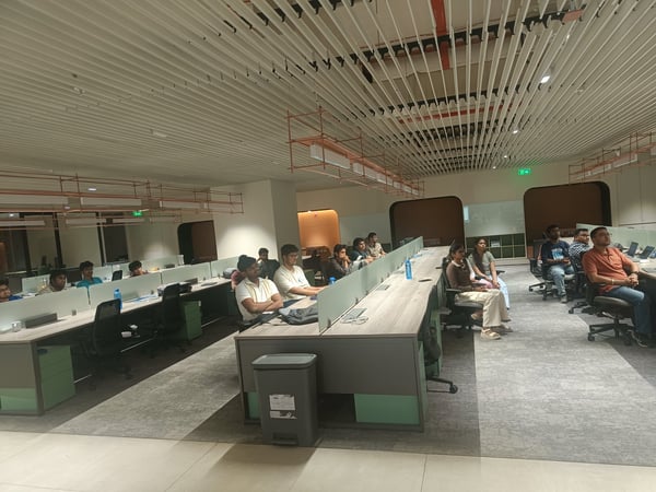 A group of people are seated in a modern office environment, appearing to attend a presentation or meeting. They are facing towards the right side of the frame in a large, well-lit room with a distinctive slatted ceiling.