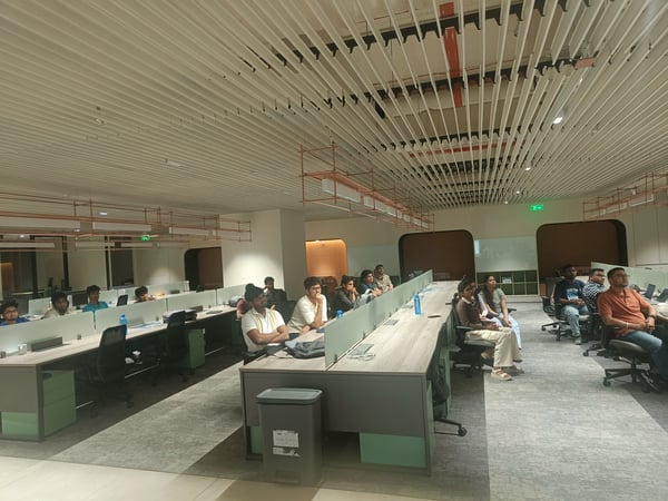 A group of people are seated at desks in a modern office environment, seemingly focused on a presentation or meeting occurring off-camera.