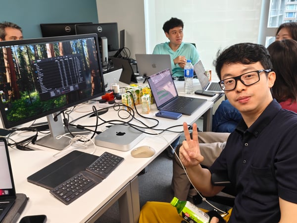 A group of people are working in a modern office environment filled with computers, laptops, and various drinks on a long white desk.