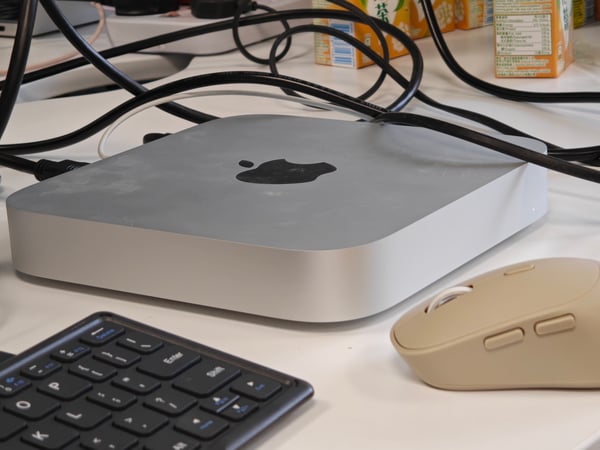 A close-up photograph of an Apple Mac Mini sitting on a white desk, surrounded by a tangle of black cables, a keyboard, and a beige computer mouse.