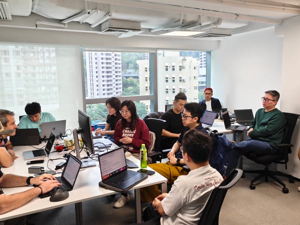 A group of people are gathered in a bright office space, working on laptops and engaged in a collaborative session. Large windows in the background provide a view of tall city buildings.