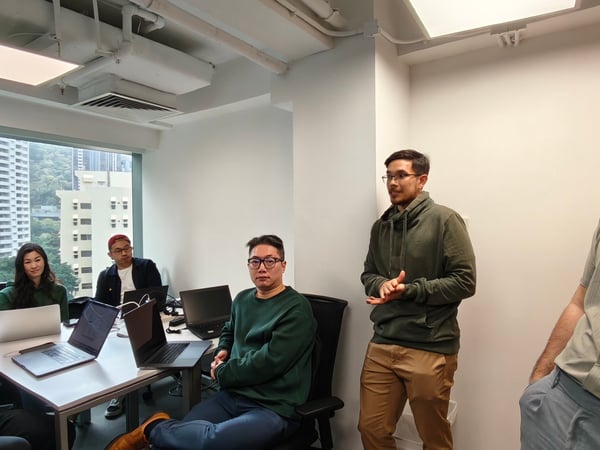 A group of people are gathered in a bright office space for a meeting or presentation, with several individuals seated around a table with laptops while one person stands and speaks.