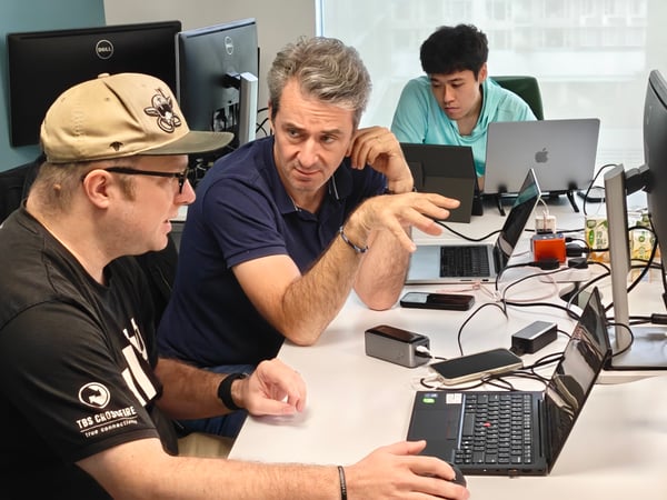 A group of people are working together in a modern office environment, with two men in the foreground engaged in a focused discussion over a laptop.