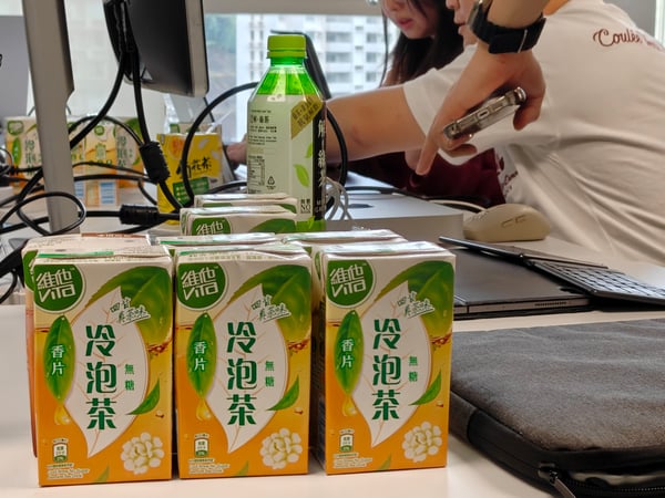 A close-up photograph of several cartons and a bottle of Vita brand cold brew tea arranged on a white office desk, with people working in the background.