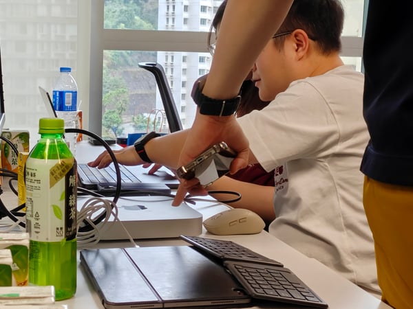 A person in a white t-shirt is working at a desk with a laptop and monitor, while another person's arm points toward a Mac Mini. The workspace is cluttered with various drink containers, including green tea cartons and a water bottle.