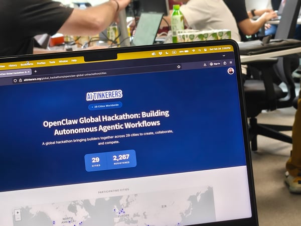 A close-up photo of a laptop screen displaying a website for the OpenClaw Global Hackathon by AI Tinkerers, with an office environment and people visible in the blurred background.