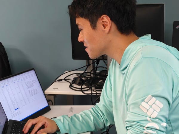A man in a light blue long-sleeved shirt is focused on a laptop screen in an office environment, with another person partially visible on the left.