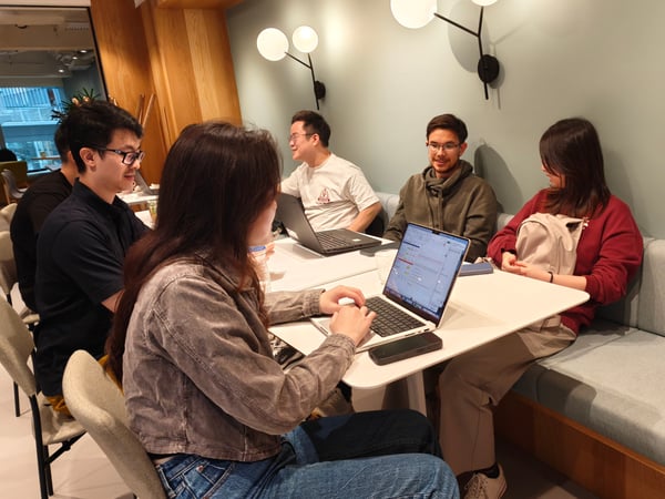 A group of young adults are sitting around a table in a modern, well-lit indoor space, with some using laptops and others engaged in conversation.
