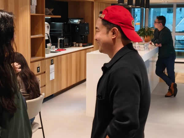 A man in a red baseball cap is smiling and talking to a woman in a green hoodie in a modern office breakroom or kitchen area.