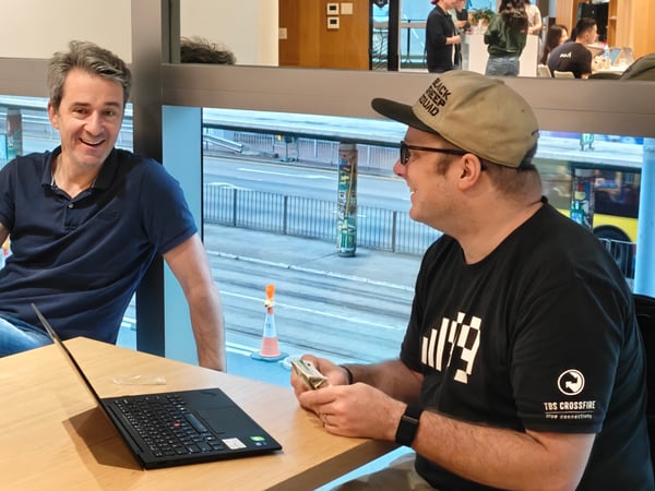 Two men are sitting at a wooden table in an indoor setting, engaged in a conversation with a laptop open between them. Large windows in the background reveal a city street with a pedestrian bridge and a bus.