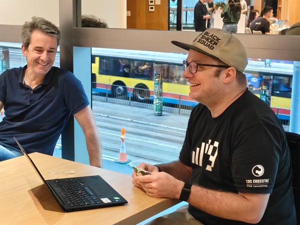 Two men are sitting at a wooden table in a brightly lit indoor space, sharing a laugh while one uses a laptop. A large window behind them reveals a city street with a passing yellow bus.