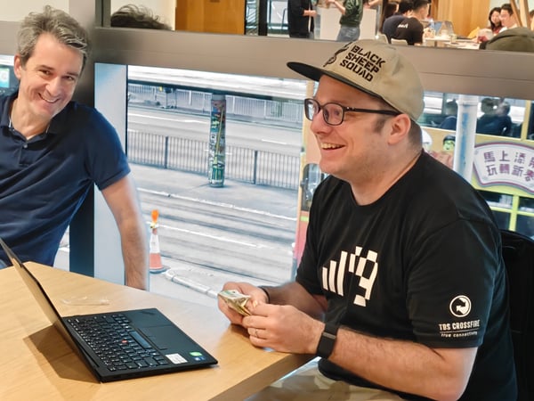 Two men are sitting at a wooden table with an open laptop, smiling and laughing in what appears to be a cafe or office with a large window overlooking a city street.