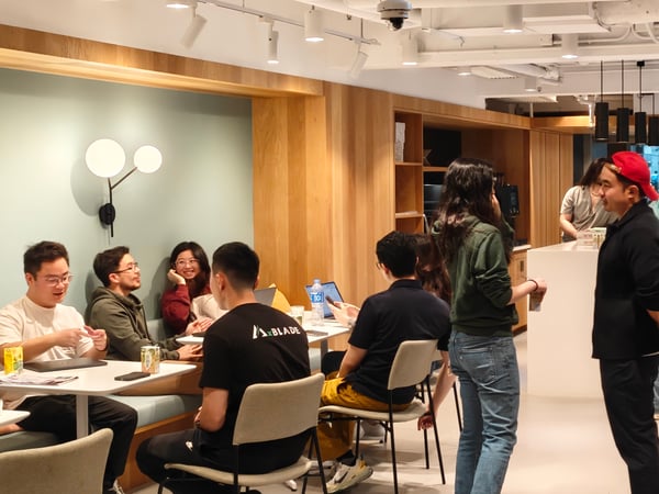 A group of individuals are gathered in a modern, well-lit indoor common area, some seated at tables with laptops and others standing and talking.