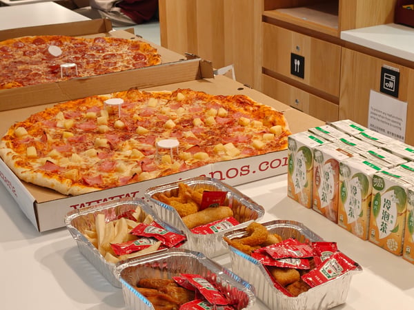 Two large pizzas, several foil containers of fried snacks with ketchup packets, and rows of tea cartons are arranged on a white table.