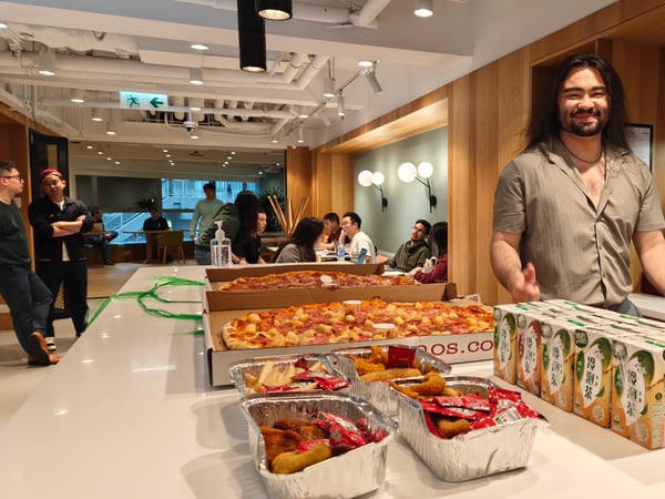 A man with long hair stands behind a kitchen counter filled with a large spread of pizzas, fried snacks, and juice boxes in a modern office setting.
