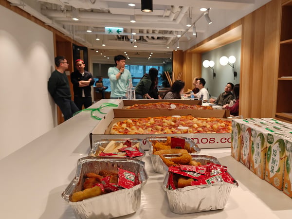 A long white table in a modern office common area is filled with party food like pizzas, fried snacks, and juice boxes, with a group of people socializing in the background.