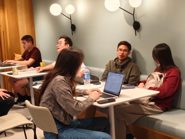 A group of young adults are sitting at tables in a modern, well-lit indoor space, possibly a cafe or co-working area, focused on their laptops and engaged in conversation.