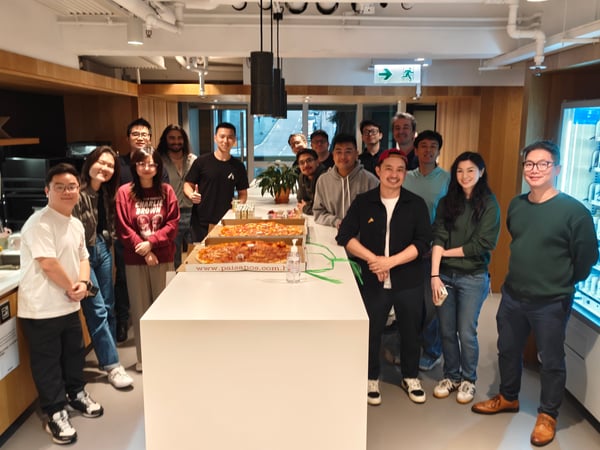 A group of colleagues posing for a photo around a table with large pizzas in a modern office pantry.