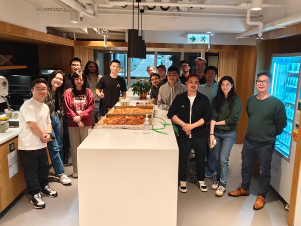 A group of people gathered around a long white table with large pizzas in a modern office pantry, posing for a group photo.