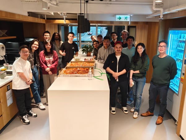A group of approximately fifteen people are gathered in a modern office pantry, posing for a photo around a white kitchen island with pizza boxes.