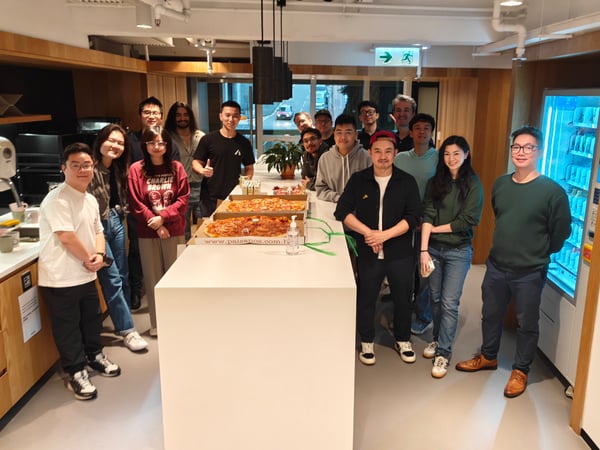 A group of colleagues posing for a photo around a kitchen island with pizza boxes in a modern office breakroom.