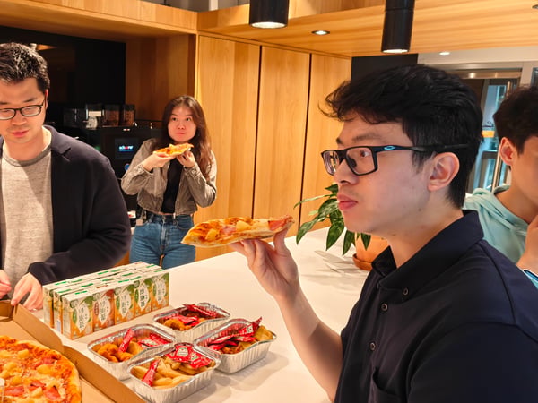 A group of people are gathered in a modern kitchen or breakroom area, enjoying a meal of pizza, fried food, and juice.