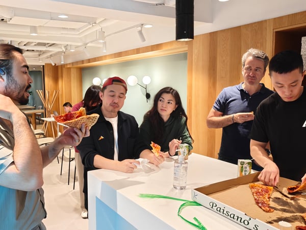 A group of people are gathered around a white counter in a modern indoor space, enjoying pizza from a Paisano's box.