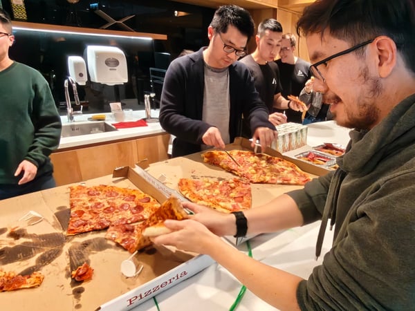 A group of men are gathered around a counter in an indoor setting, sharing and eating large pizzas from open boxes.