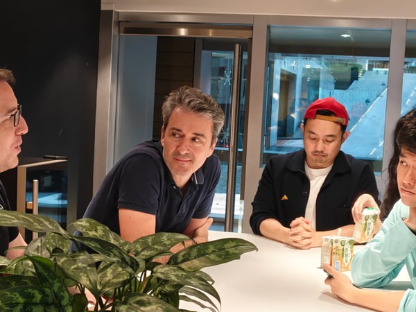 Four men are seated around a white table in an indoor setting, engaged in a conversation with a large potted plant in the foreground.