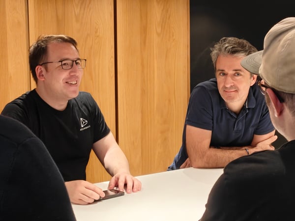 A group of men are gathered around a white table in an indoor setting with wooden wall panels, appearing to be in a meeting or discussion.