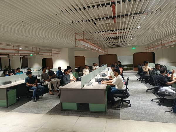 A group of people are seated at long rows of desks in a modern office space, all looking towards the left side of the room as if attending a presentation.