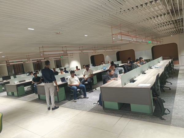 A group of people are gathered in a modern, open-plan office. One man is standing and appears to be speaking to others who are seated at long, shared desks with laptops.