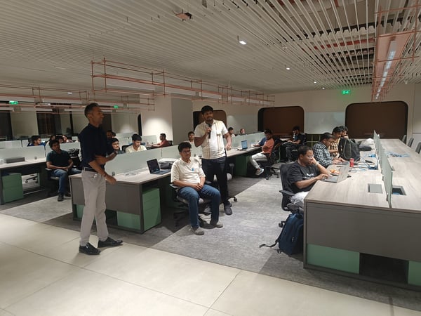 A group of people are gathered in a modern office space, with one man standing and speaking into a microphone while others sit at their desks with laptops.
