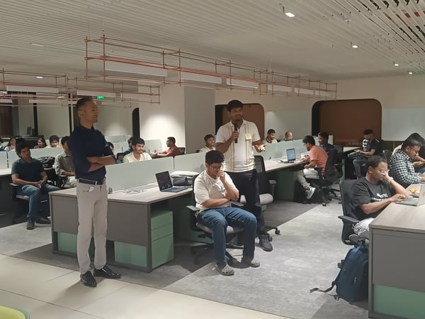 A group of people are gathered in a modern office or coworking space for a presentation or demo. One man is standing and speaking into a microphone while others sit at desks with laptops, listening.