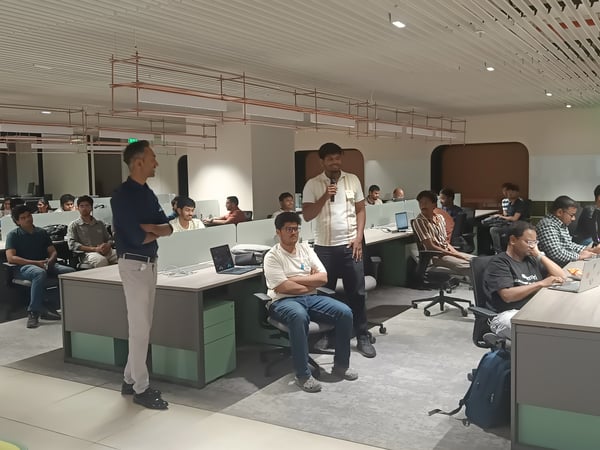 A group of people in a modern office environment, where one man is standing and speaking into a microphone while others listen from their desks.